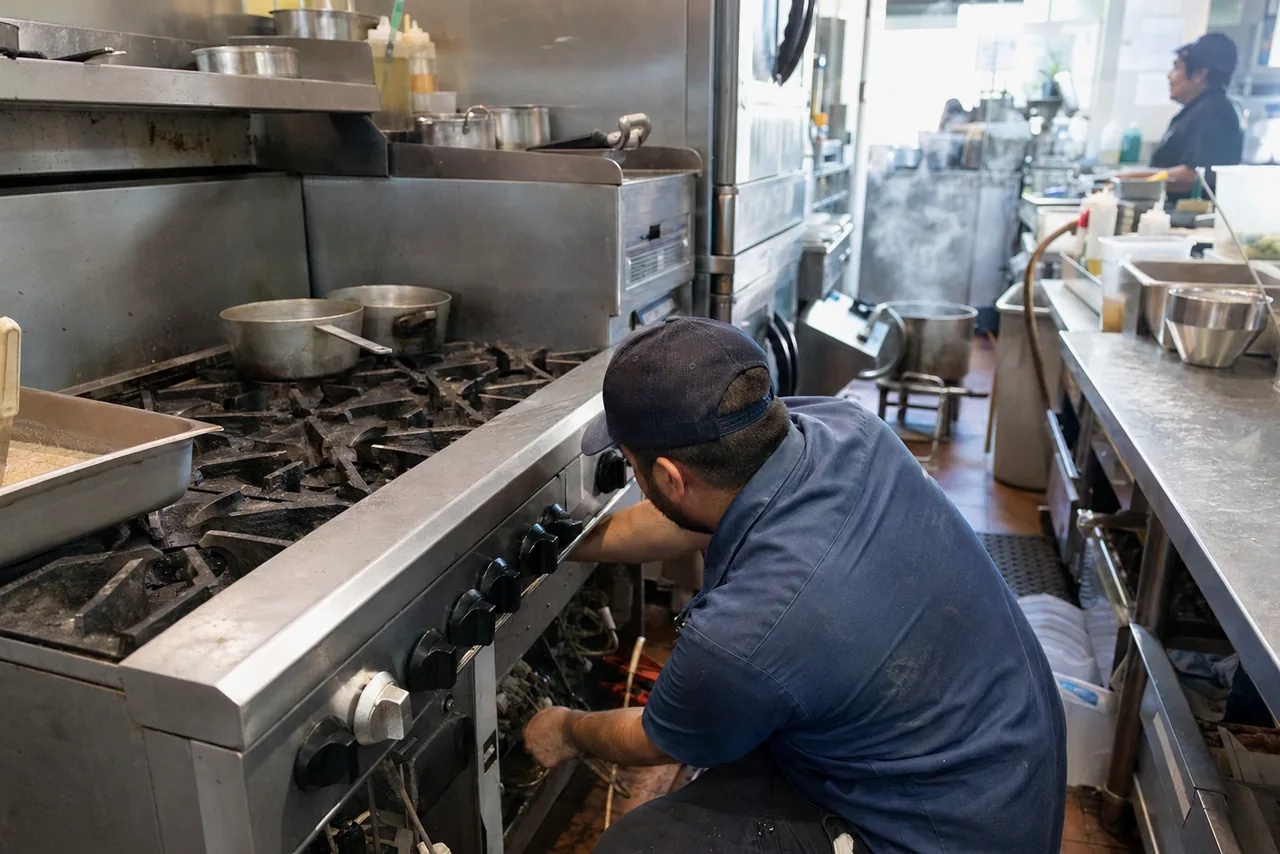 A technician kneeling at the front of a stainless gas range with the fascia off, the burner tops loaded and a colleague working further down the line