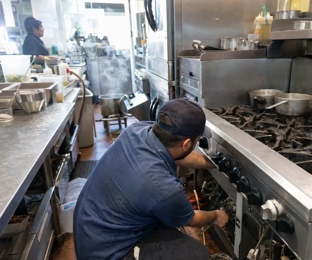 A technician working at the control fascia of a stainless gas range, the wiring behind the knob row exposed in a busy kitchen