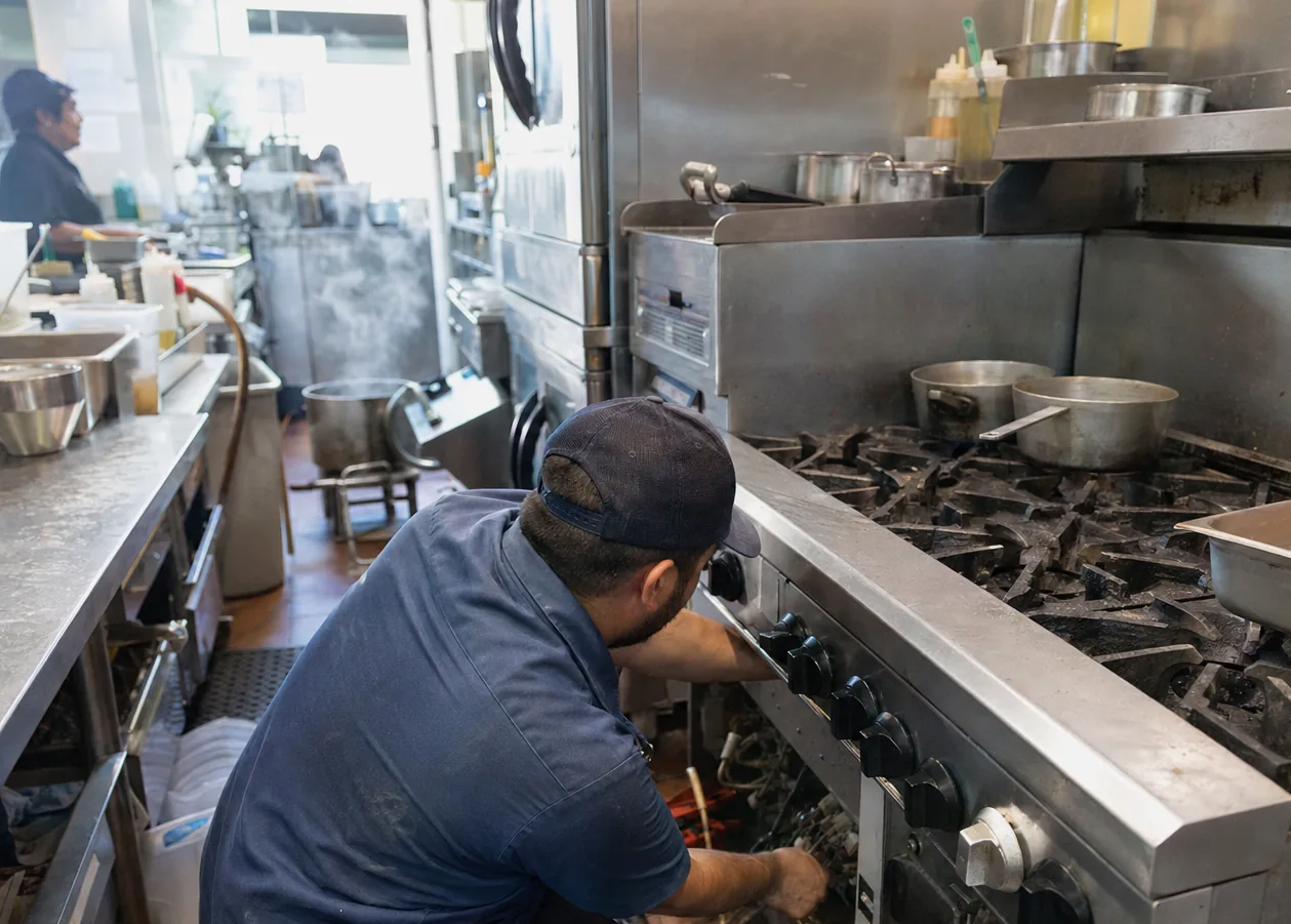 A technician working at floor level in front of a stainless appliance in a narrow kitchen aisle, panel off and the drain run exposed