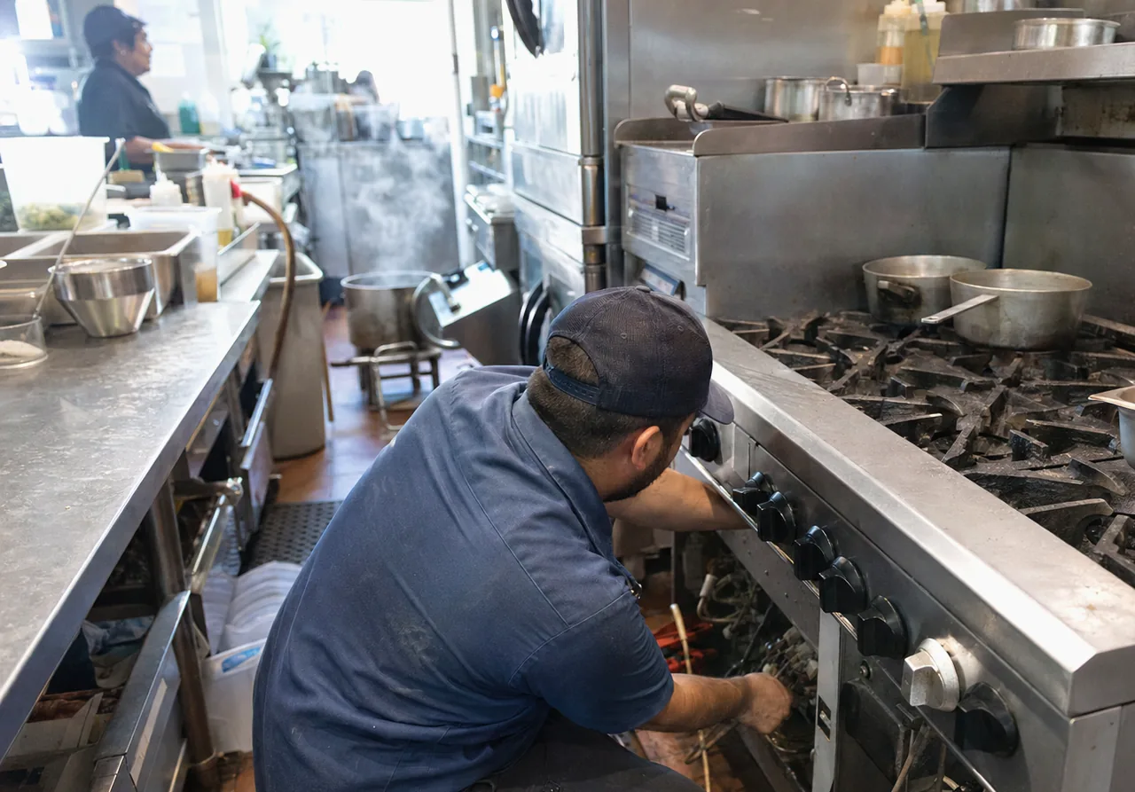 A technician on one knee at a tall stainless cooking appliance, its lower access panel off and the element wiring reachable inside